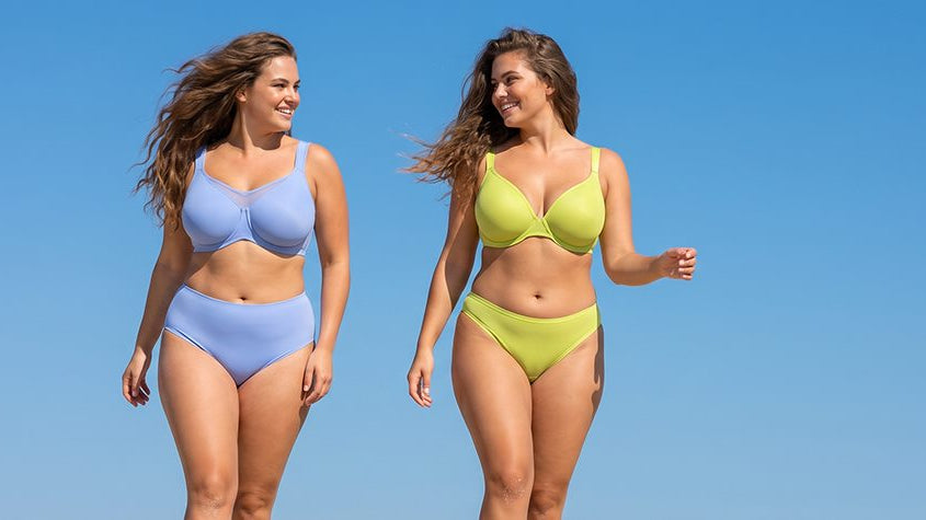 Two plus-size models walking outdoors under blue sky — one in periwinkle CoolComfort unlined minimizer bra, one in chartreuse Joan unlined t-shirt bra, both with matching bottoms