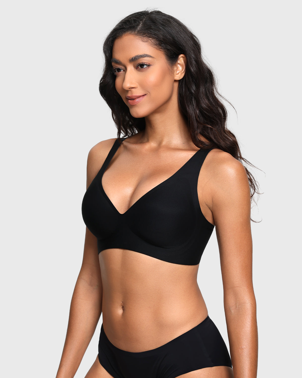A woman modeling a black two-piece lingerie set consisting of a bra and matching underwear, photographed against a solid light gray background.