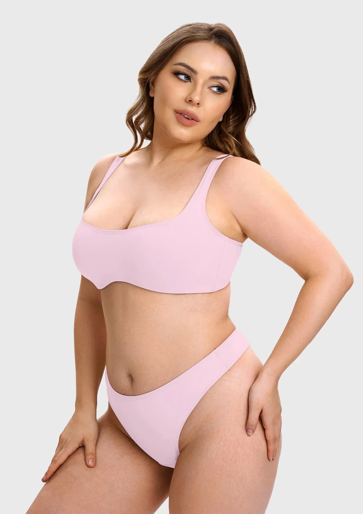 A plus-size model wearing a light pink two-piece bikini set, consisting of a scoop-neck bralette and matching high-cut bottoms, against a solid light gray background.