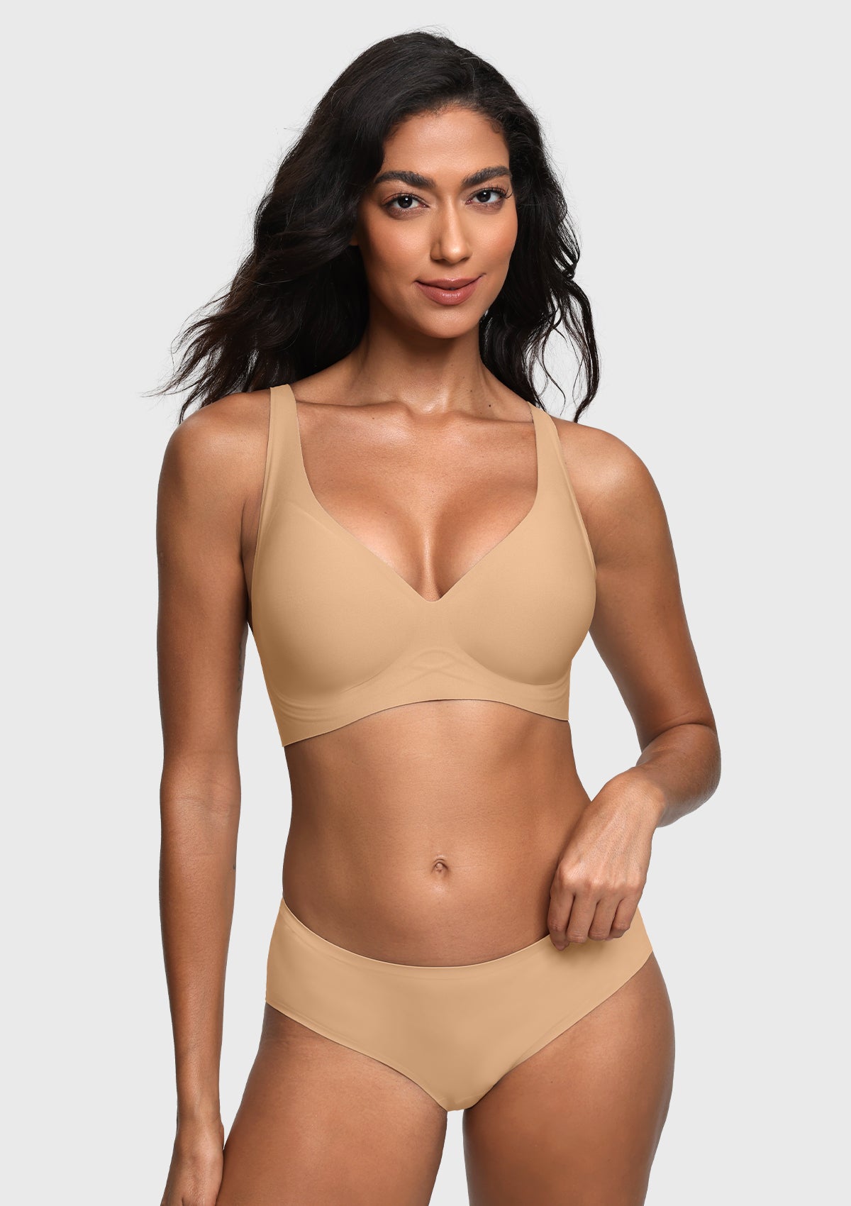 A woman modeling a light beige wireless bra and matching high-waisted briefs against a light gray studio background.